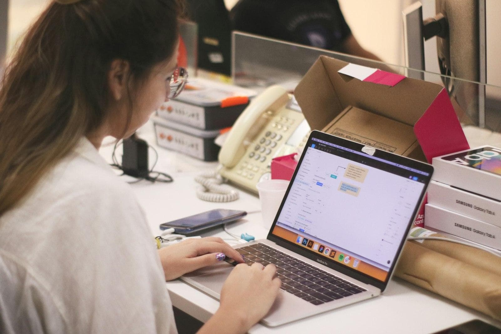 A shop worker answering messages on a laptop at the counter, between the desk phone and boxes waiting to ship
