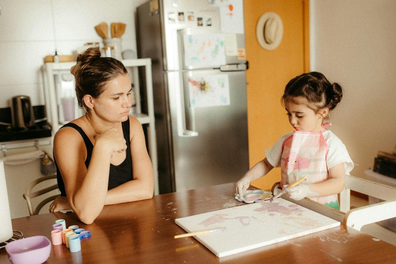 A parent watching a young child paint at the kitchen table, kids’ drawings on the fridge behind them