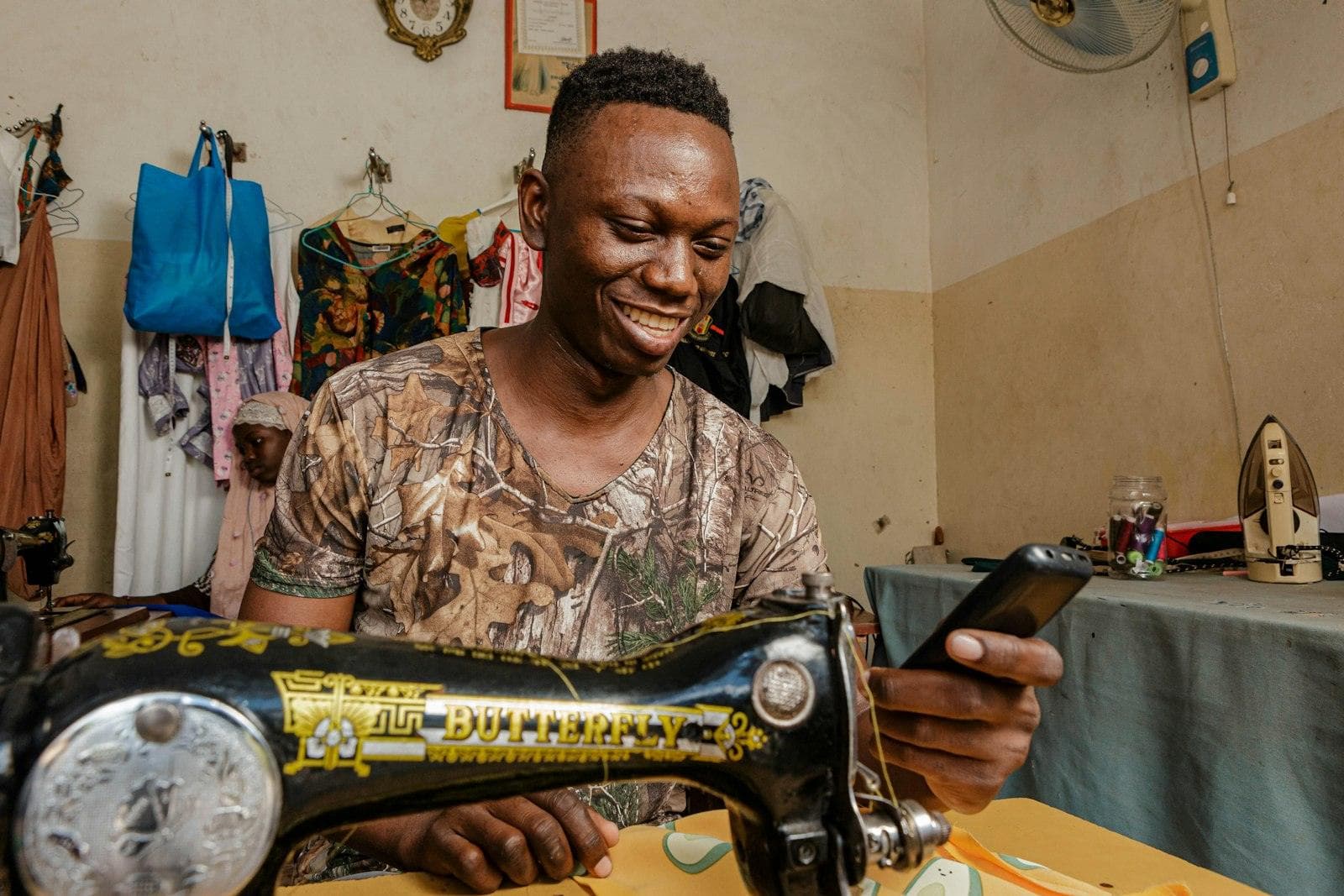 A tailor smiling at a phone at the sewing machine in a small workshop